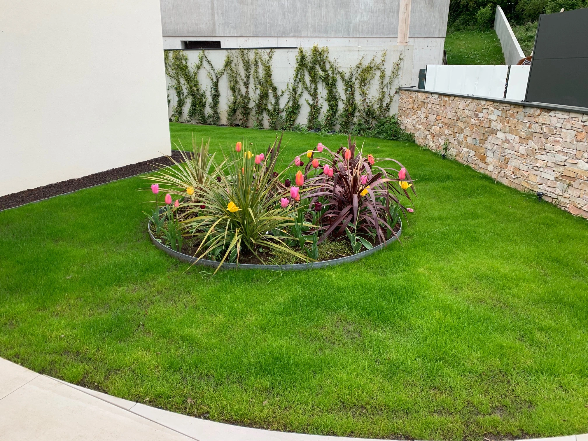 Jardin réalisé en partie au dessus d'un garage. Des dalles légères sur plots et un gazon synthétique ont été posé ainsi que des massifs de plantes fleuries. Des jasmins ont été plantés le long du murs mitoyen sur la partie pleine terre.